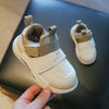 A hand holds a white kids' everyday sneaker with tan accents and gray velcro straps on a dark surface.