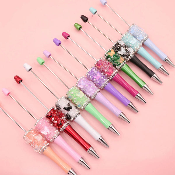 A collection of decorative rhinestone bead ballpoint pens in various colors and finishes arranged diagonally on a pink backgr