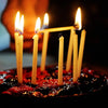 A plate of red cake decorated with lit natural beeswax taper candles in a dark setting.
