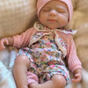 A newborn baby doll wearing a pink knitted hat and floral outfit, lying on a light brown fabric.