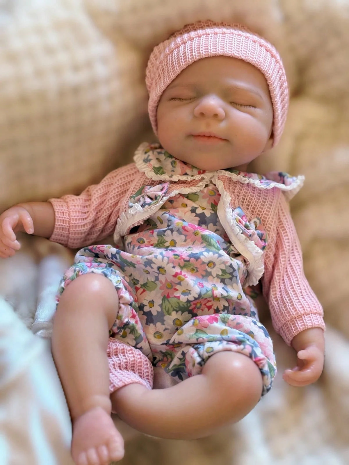A newborn baby doll wearing a pink knitted hat and floral outfit, lying on a light brown fabric.