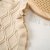 A beige knit sweater vest with a diamond pattern and ruffled edge, displayed next to a wooden round object.