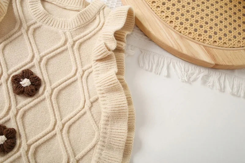 A beige knit sweater vest with a diamond pattern and ruffled edge, displayed next to a wooden round object.