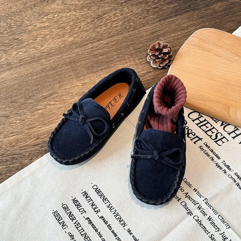 A pair of navy blue girls' moccasins with bow details resting on a piece of paper.