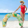 A woman stands on a beach holding an inflatable U-shaped swim ring with green and yellow tropical leaf pattern.