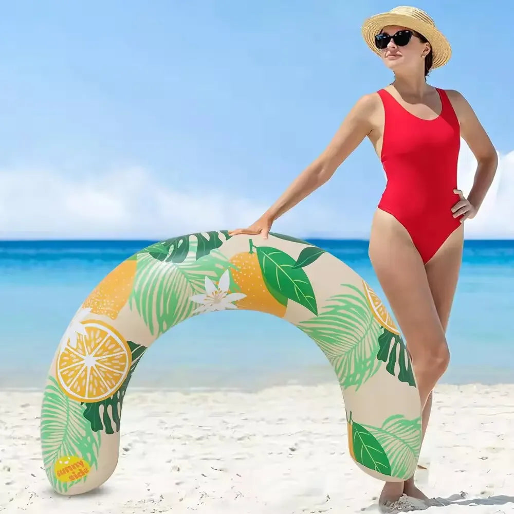 A woman stands on a beach holding an inflatable U-shaped swim ring with green and yellow tropical leaf pattern.