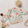 A white feeding bib with a dinosaur and animal print featuring pink, green, and yellow colors, resting on a light fabric.