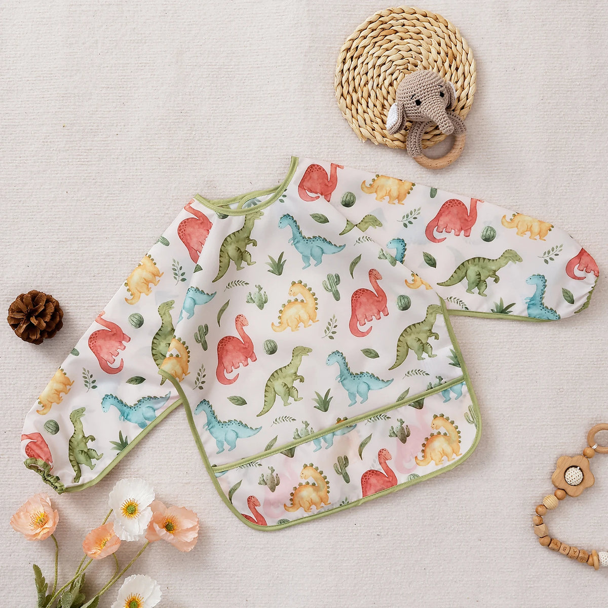 A white feeding bib with a dinosaur and animal print featuring pink, green, and yellow colors, resting on a light fabric.