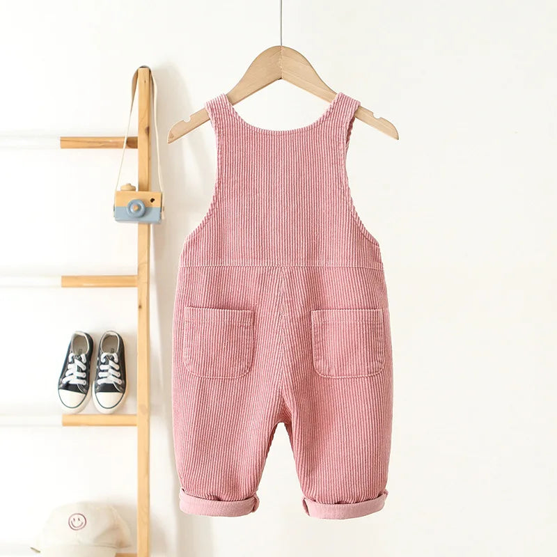 A pink corduroy toddler overalls hanging on a wooden hanger against a white wall.