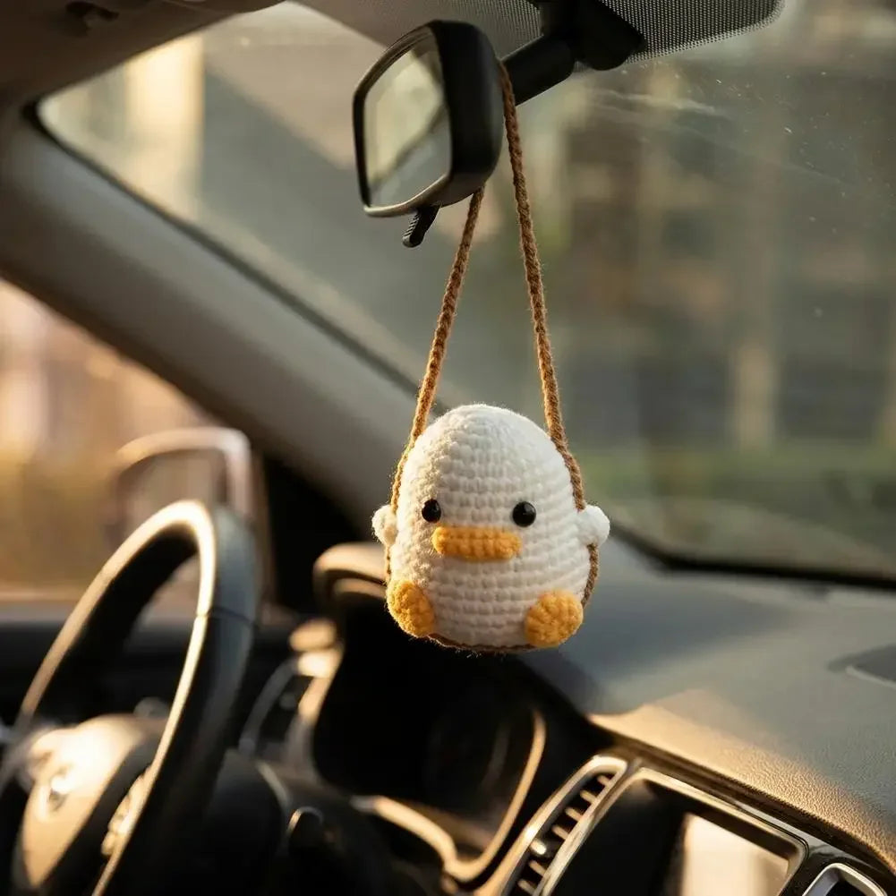 A handmade crochet white duck ornament is hanging from a rearview mirror on a car windshield.