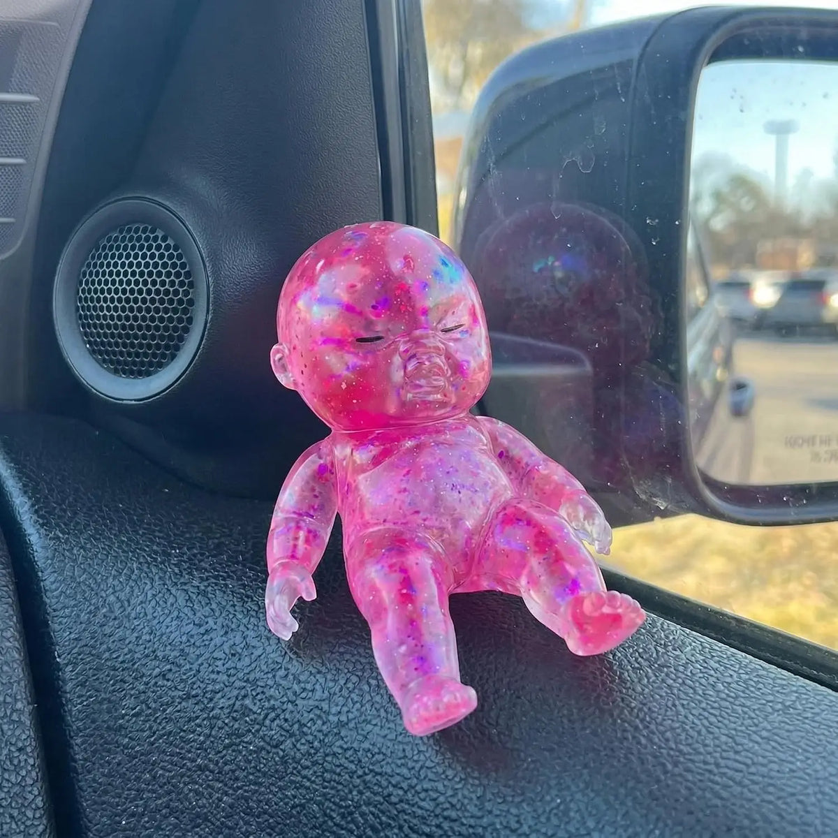 A small, pink glitter silicone squishy toy resting on a car interior panel next to a vent and a rearview mirror.