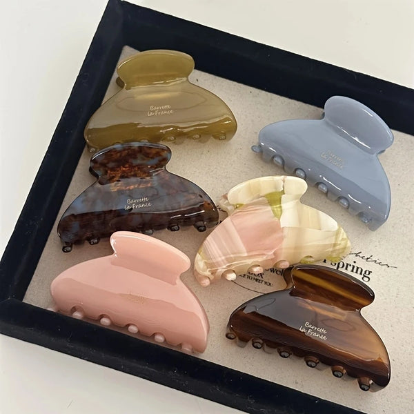 A collection of elegant acetate hair claw clips in various shades of olive green, brown, and pink displayed in a black tray.