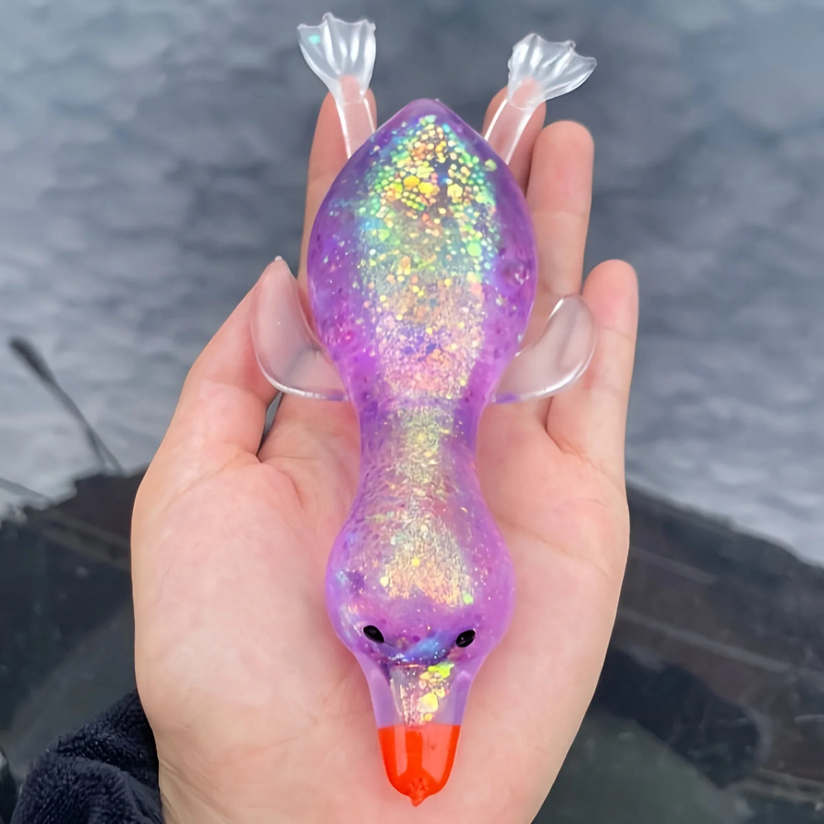 A hand holds a vibrant purple squishy duck fidget toy with iridescent glitter and an orange beak.