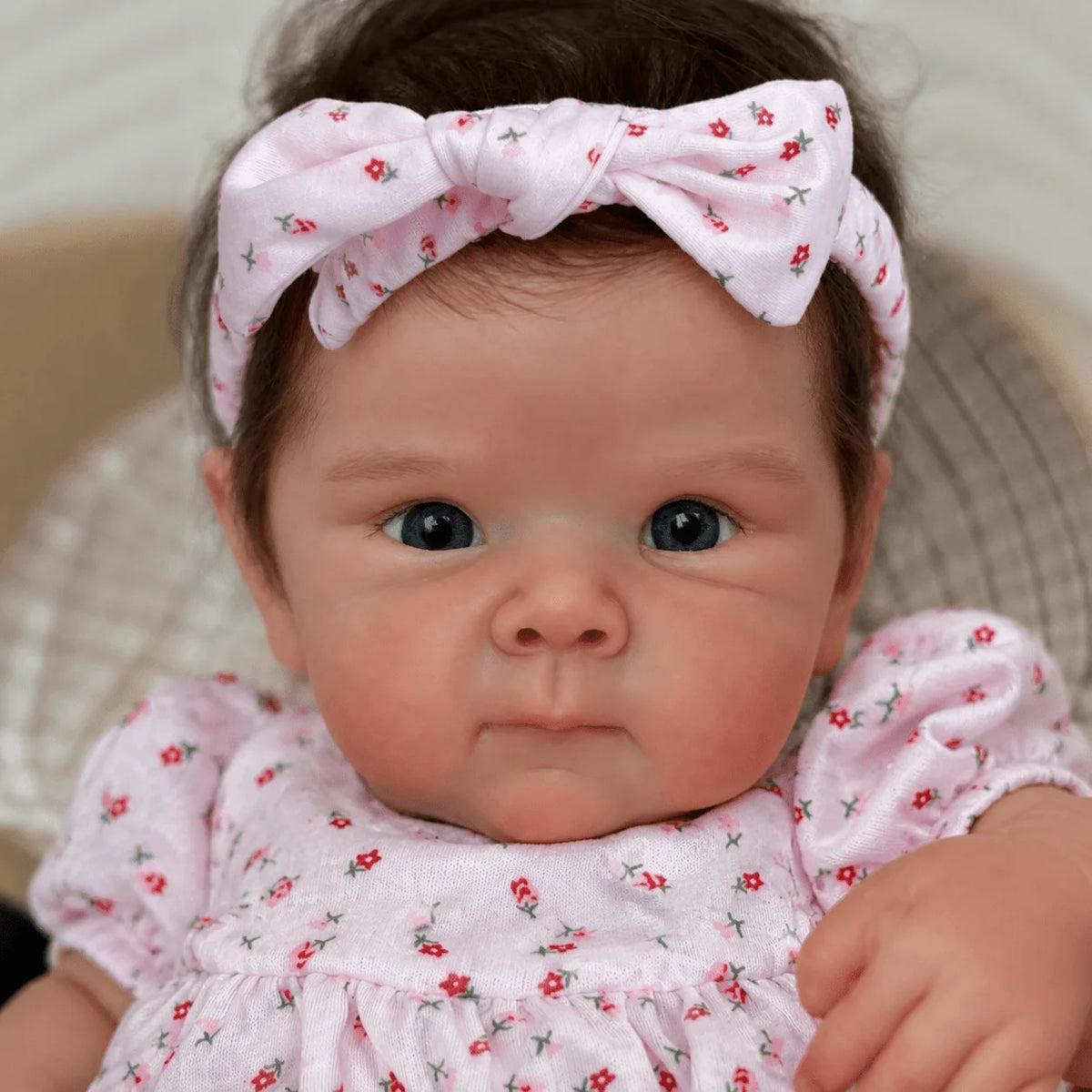 A close-up photo of a lifelike vinyl reborn baby doll with hand-rooted hair wearing a pink floral dress and bow.