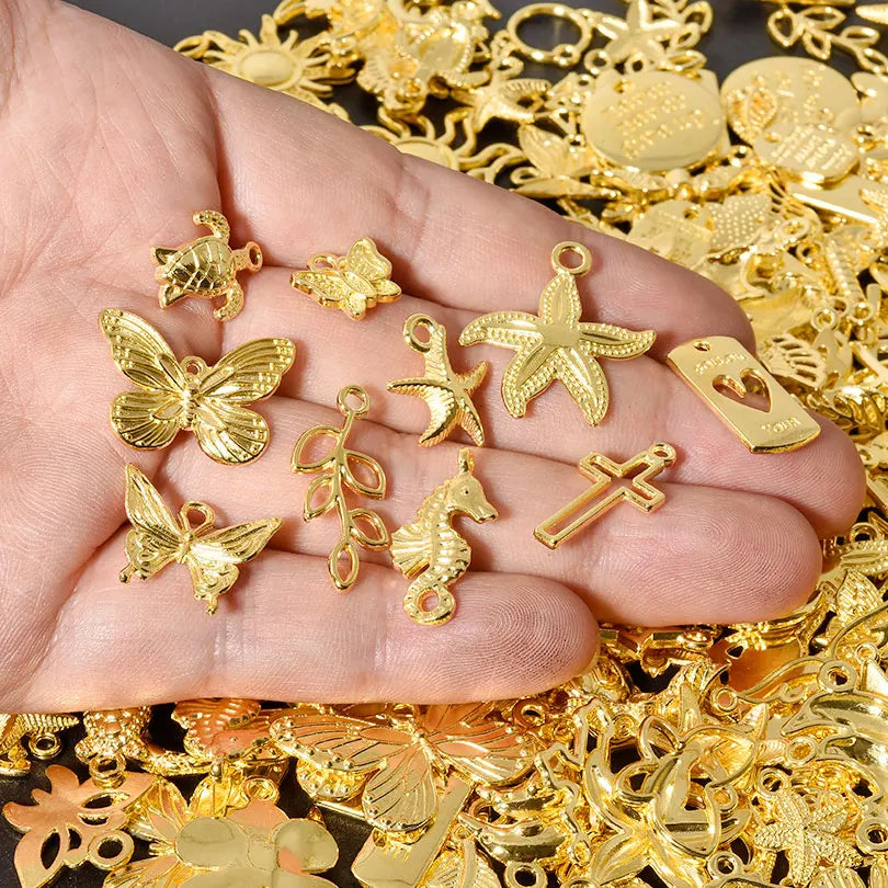 A hand holds various gold-colored mixed metal charms including a butterfly, star, and cross for DIY jewelry making.