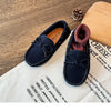A pair of navy blue girls' moccasins with a bow detail resting on a white cloth next to a wooden block and a pinecone.