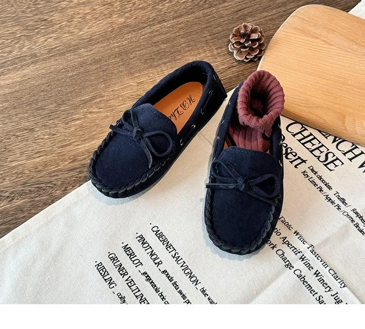 A pair of navy blue girls' moccasins with a bow detail resting on a white cloth next to a wooden block and a pinecone.