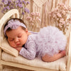 A newborn baby wearing a purple lace headband and pink tutu dress is resting on a light wooden surface.
