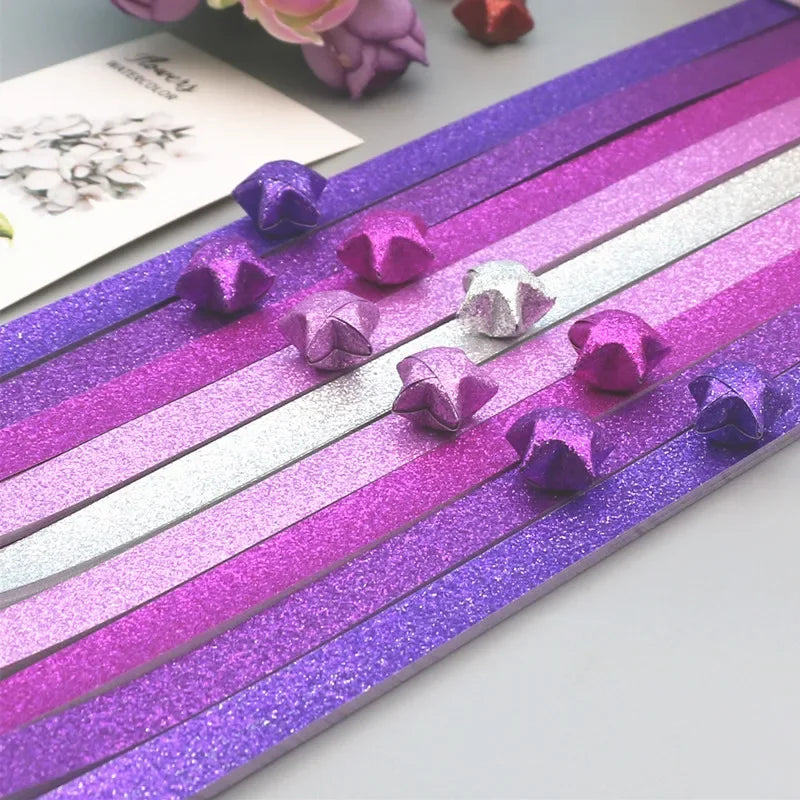 A collection of glitter origami star paper strips in purple and silver shades laid out on a surface.