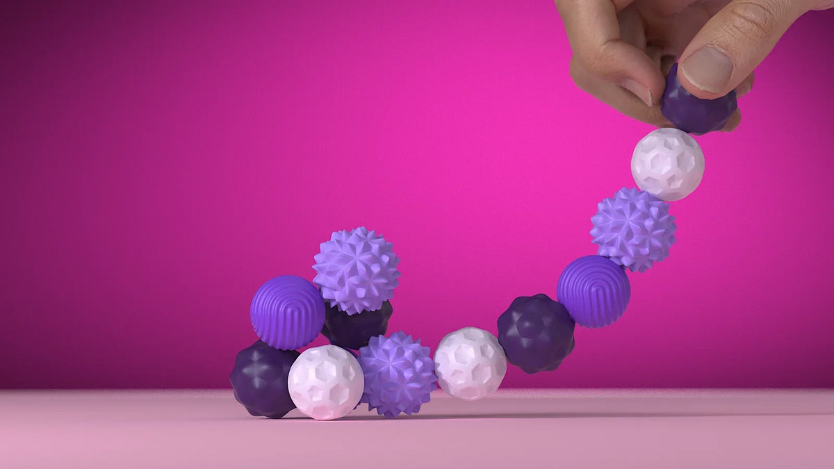 A hand is holding and arranging purple and white soft sensory silicone massage balls on a pink surface.