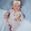A newborn reborn baby doll wearing a pink outfit and a cream bow, lying on white bedding.