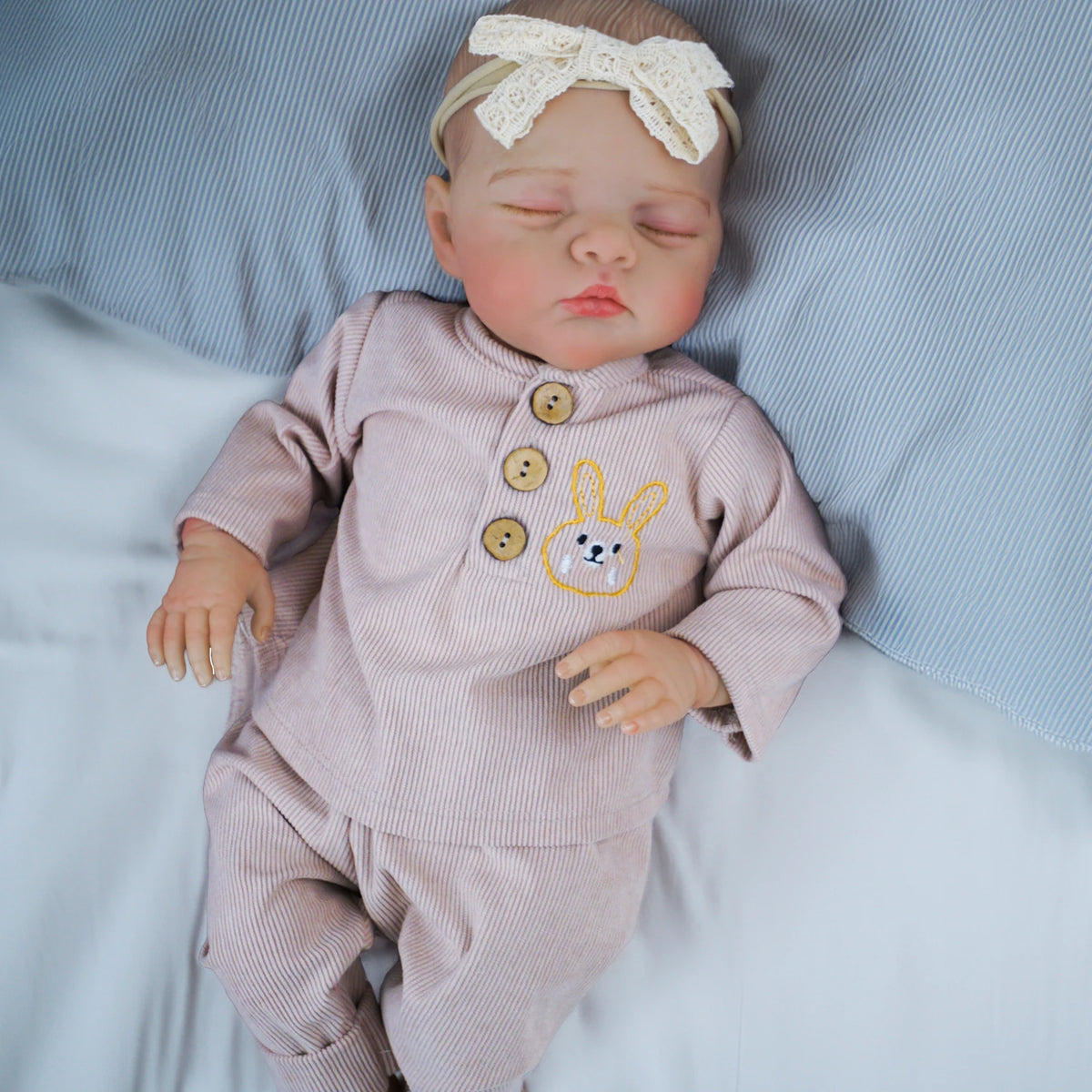 A newborn reborn baby doll wearing a pink outfit and a cream bow, lying on white bedding.