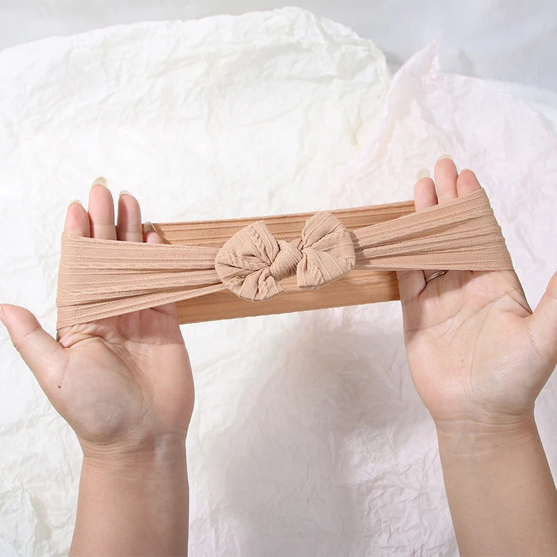 Three soft beige stretch baby headbands with bows are held in open hands against a white background.