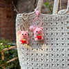 A pink crocheted mini pig keychain with a gold-toned ring hanging from a natural fiber tote bag.