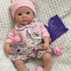 A realistic soft silicone reborn baby doll wearing a pink knitted hat and floral outfit, lying on a white fuzzy surface.