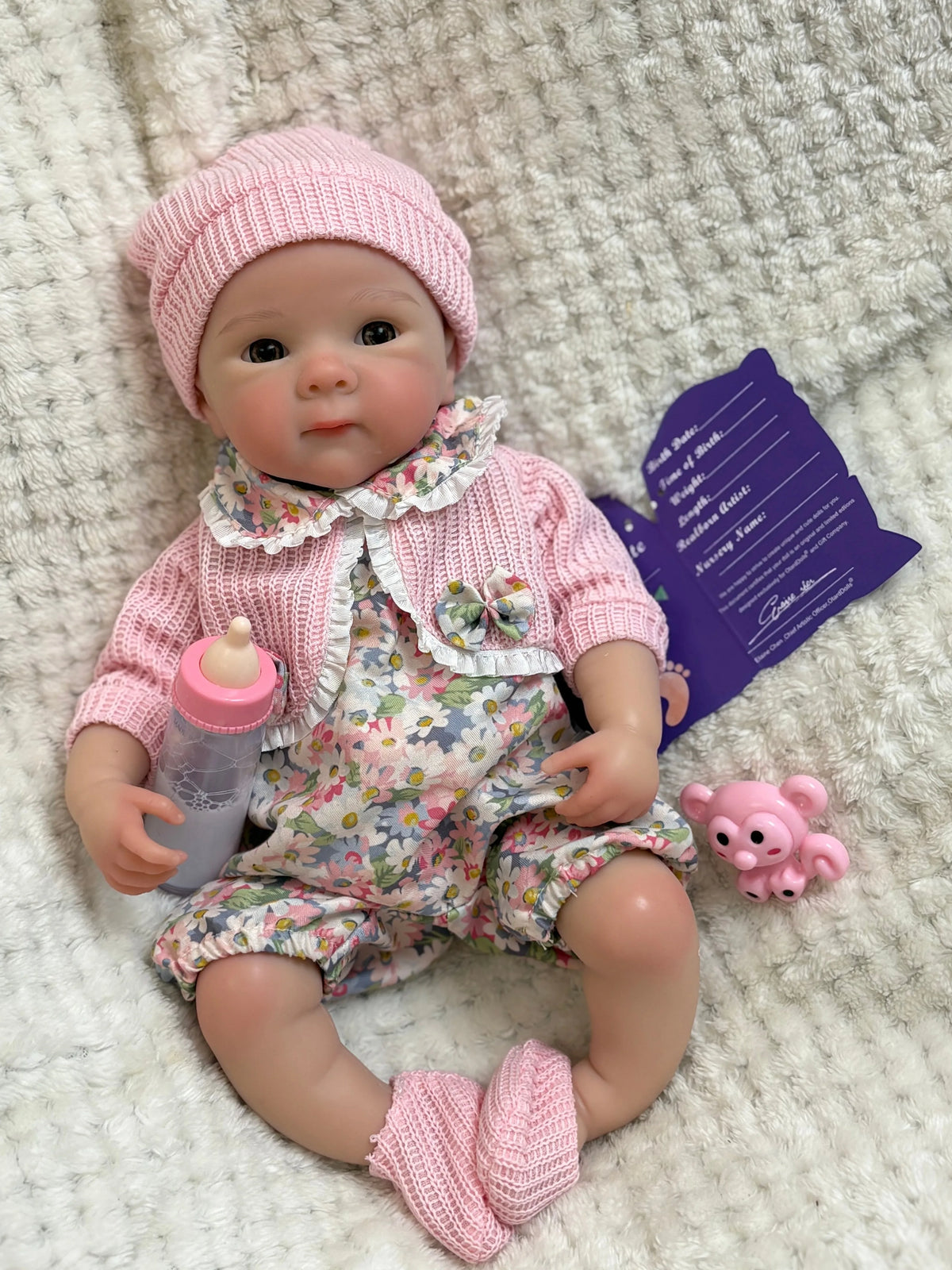 A realistic soft silicone reborn baby doll wearing a pink knitted hat and floral outfit, lying on a white fuzzy surface.