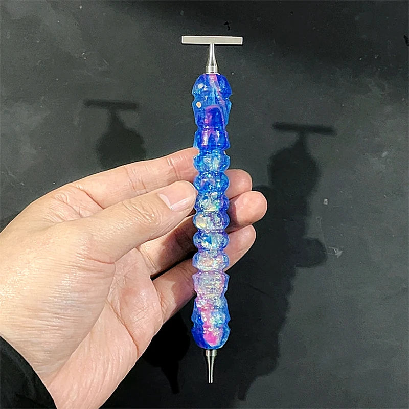 A hand holds a colorful diamond painting pen with a stainless steel tip, displaying iridescent blue and purple hues.