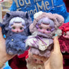 A hand holds a Skullpanda You Found Me! plush doll figure with purple hair and pink accents against a blue background.
