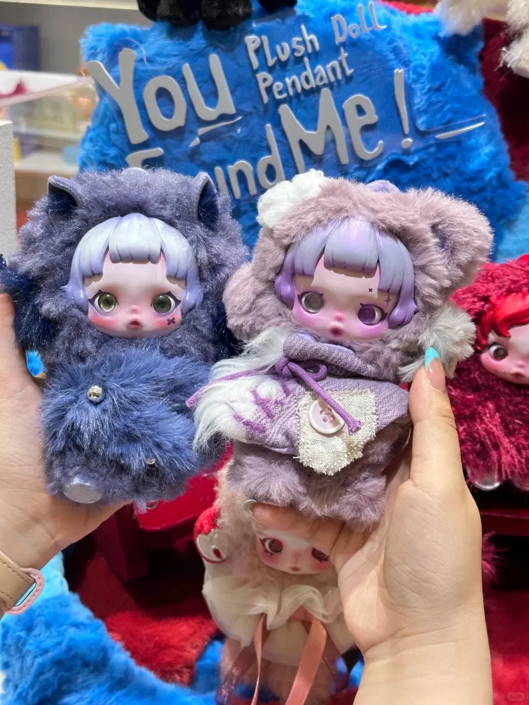 A hand holds a Skullpanda You Found Me! plush doll figure with purple hair and pink accents against a blue background.