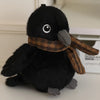 A black cartoon bird plush with a brown plaid scarf resting on a white surface.