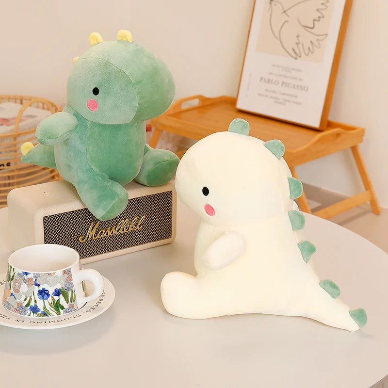 A soft green dinosaur plush toy sitting next to a white dinosaur plush toy on a table.