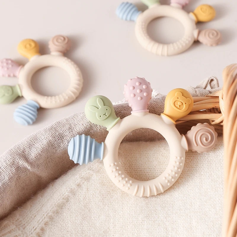 Eine Sammlung von Silikon-Teether-Ringen in verschiedenen Pastellfarben, darunter rosa, gelb und grün, auf beige Stoff