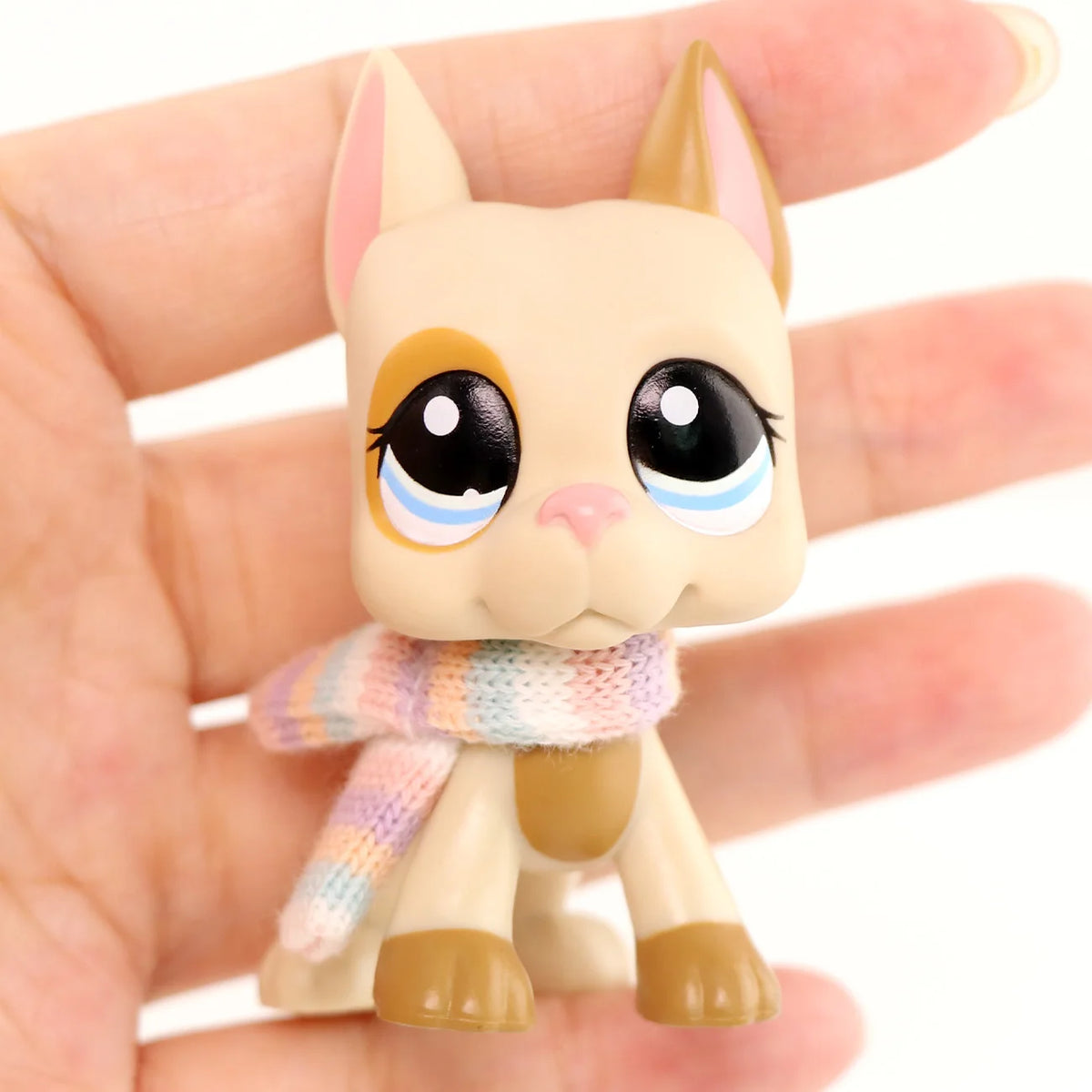 A small, light beige cat figure wearing a multicolored knitted scarf is held in a person's hand.