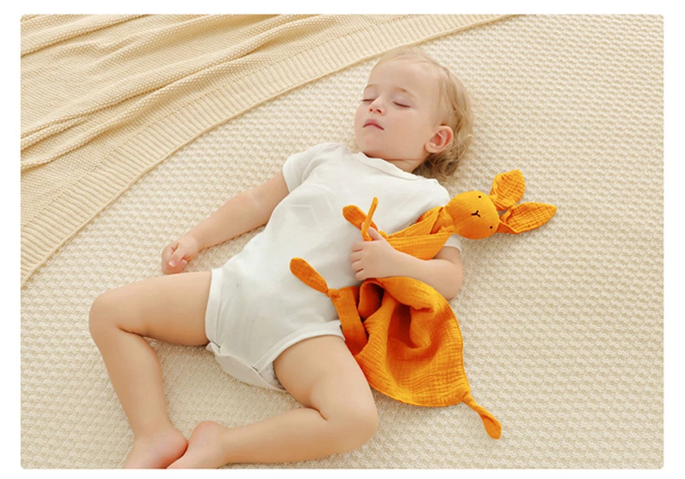 A baby lies on a cream blanket, gently holding an orange fabric toy.