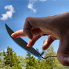 A hand holds a black and blue mini finger surfboard skater toy against a bright blue sky with green trees in the background.