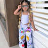 A girl wearing a white two-piece outfit with a suspender top and long pants, sunglasses, and braids, standing next to a white