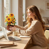 A young woman is assembling a DIY rose and sunflower building block bouquet at a wooden table near a window.