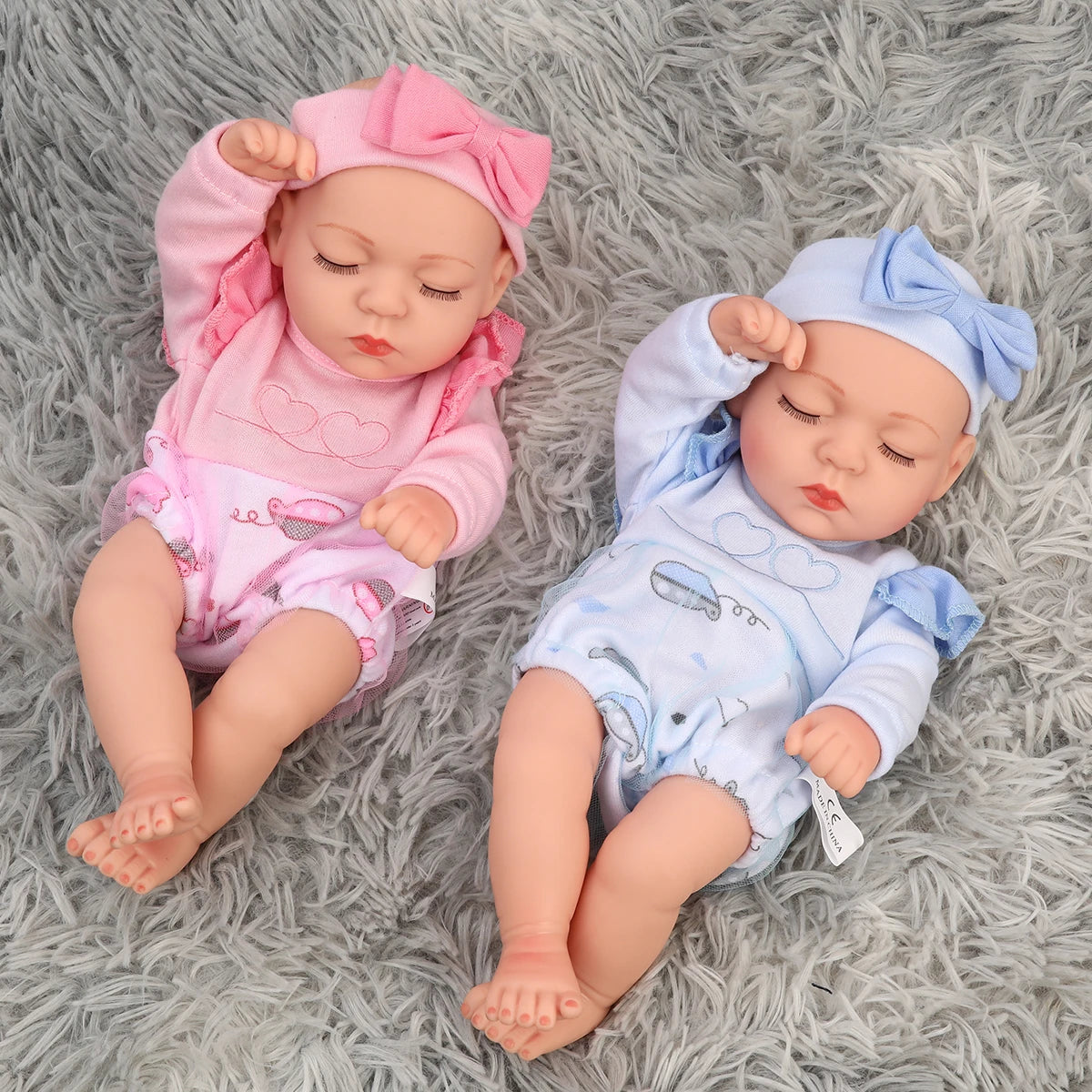 A pair of lifelike soft vinyl reborn dolls with pink and blue outfits lying on a gray fluffy surface.