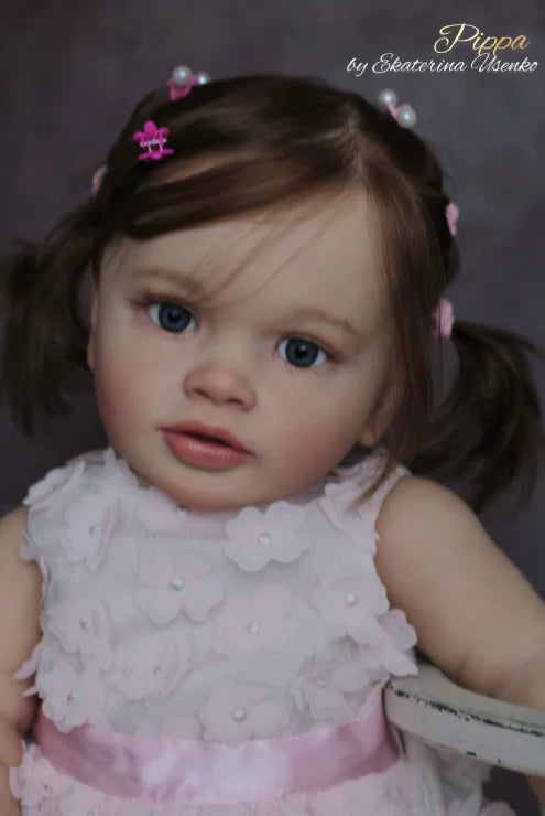 A close-up photo of a lifelike reborn baby doll with brown hair and blue eyes wearing a pink floral dress.