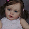 A close-up photo of a lifelike reborn baby doll with brown hair and blue eyes wearing a pink floral dress.