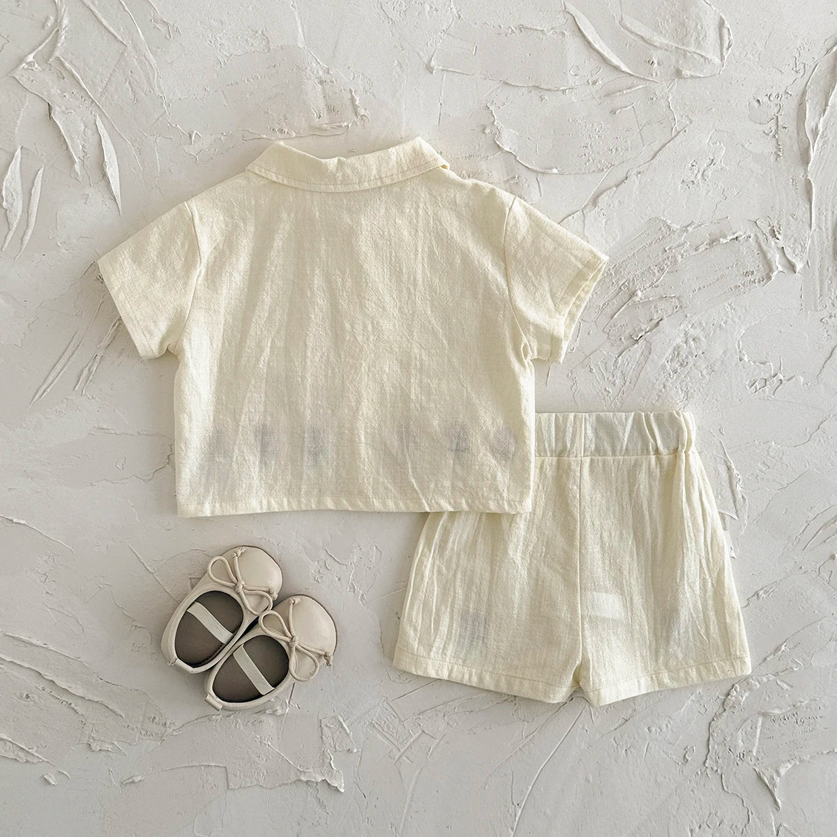 A cream-colored baby cotton shirt and shorts set with a subtle flower pattern laid on a white surface.