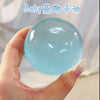 A hand holds a clear blue glass squeeze ball, likely made of coconut oil, against a light background.