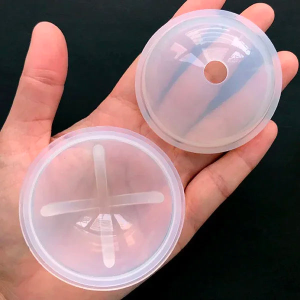 A hand holds two clear silicone molds for resin crafting, showing a circular mold with a hole and a cross-shaped mold.