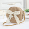 A handwoven straw crossbody bag with a beige lace bow resting on a white surface.