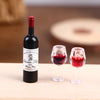 A black miniature resin wine bottle and two corresponding tasting glasses are displayed on a wooden surface.