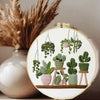 A cross stitch kit featuring green potted plants hanging from strings in a circular embroidery design.